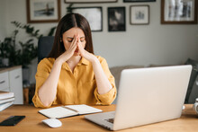 Female Freelance With Eyes Closed Is Massaging Head Temples With Suffering Face Expression. Tired And Worn Out From Online Work Young Woman Sitting In Front Laptop Holding Head