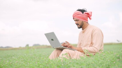 Wall Mural - Young indian farmer busy wrking on laptop while sitting at agricultural farmland - concept of technology, digital communication and modern farming.