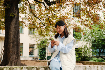 Wall Mural - Young asian girl using mobile phone while sitting outdoors