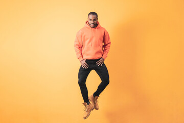 A dark-skinned man in an orange hoodie looks at the camera as he jumps in front of an orange wall.