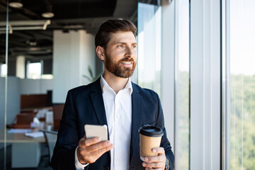 Confident middle aged businessman in formal wear with coffee to go and smartphone standing near window in office