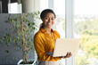 © Prostock-studio - Portrait of happy businesswoman using laptop in office interior, standing near window and smiling at camera