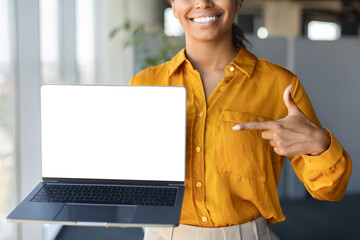 Female black entrepreneur pointing at laptop computer with blank screen for website design, standing in office, mockup