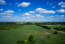 An Aerial View Of A Green Field With Trees And A Blue Sky With White Clouds Above It And A Road Running Through The Center Of The Field, And A Few Trees To The Left. .