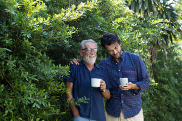 Wall Mural - Happy senior man and adult man walking and drinking coffee or tea outdoor garden. Two happy men spending time together in the park. Happy family, holiday and people concept