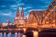Scenic view of illuminated bridge over Rhine river whith trains and tourists passing by and the Cologne Cathedral in the evening at the blue hour.