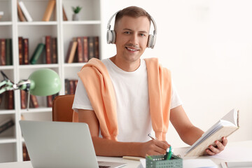 Wall Mural - Young man with headphones, book and laptop studying online at home