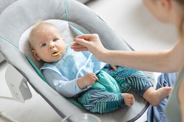 Wall Mural - Mother spoon feeding her baby boy child in baby chair with fruit puree. Baby solid food introduction concept