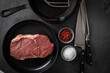 Entrecote Steak roh vom Rind mit Meersalz und roter Pfeffer auf Teller bei Vorbereitung in Küche mit Messer, Fleischgabel, Gusseisern Pfanne und Pfannenwender auf Schiefer Stein Hintergrund Schwarz