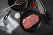 Entrecote Steak roh vom Rind mit Meersalz und roter Pfeffer auf Teller bei Vorbereitung in Küche mit Messer, Fleischgabel, Gusseisern Pfanne, Plastik Vakuumbeutel und Pfannenwender auf Schiefer Stein 