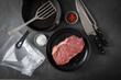 Entrecote Steak roh vom Rind mit Meersalz und roter Pfeffer auf Teller bei Vorbereitung in Küche mit Messer, Fleischgabel, Gusseisern Pfanne, Plastik Vakuumbeutel und Pfannenwender auf Schiefer Stein 