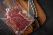 Entrecote Steak roh vom Rind in Plastik Vakuumbeutel für Vakuum marinieren und Sous Vide garen mit Meersalz auf Holzbrett in Küche mit Messer, Fleischgabel und Schiefer Stein Hintergrund Schwarz