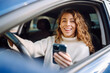 © maxbelchenko - Beautiful young woman is sitting inside a car on the drivers seat with phone. Business, taxi, technologie, internet, online concert.