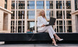 © Iona - Upset young blonde entrepreneur woman using laptop with sad facial expression  sitting outdoors against large building. Pretty student girl preparing for exam. Overloaded businesswoman typing letter.