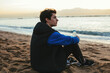 © Cavan Images - Side view of thoughtful teenage boy holding water bottle while sitting at beach