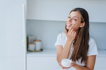 Pretty Italian young woman with long loose hair standing at living room puts skin cream on face broad smiles satisfied by skin care products and healthy lifestyle. Cosmetics. Satisfied female at home.