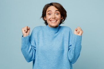 Wall Mural - Young fun caucasian woman wear knitted sweater look camera doing winner gesture celebrate clenching fists say yes isolated on plain pastel light blue cyan background studio. People lifestyle concept.