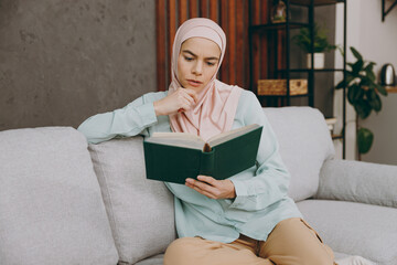 Young smart muslim woman wearing hijab casual clothes reading book sits on sofa couch stay at home flat rest relax spend free spare time in living room indoor. People middle eastern uae islam concept.