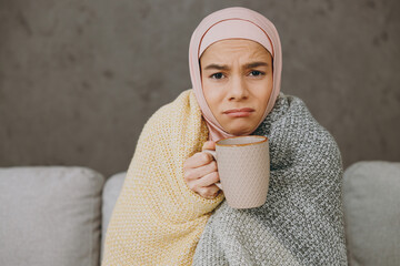 Wall Mural - Young sick muslim woman wear hijab casual clothes plaid drink tea sits on sofa couch stay at home flat rest relax spend free spare time in living room indoor People middle eastern uae islam concept.