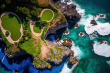 Aerial View Of A Stunning Golf Course On A Lush, Rocky Clifftop In A Posh Tourist Destination On Indonesia's Paradise Island Of Bali. In The Pristine Blue Water, Swimmers And Surfers Alike Are Active