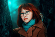 A close-up portrait of a beautiful Asian red-haired woman in warm clothes in a dark forest near tree trunks in winter.