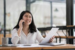 © wichayada - Happy smiling asian business woman working on laptop at office, using smart phone. Businesswoman sitting at her working place