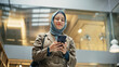 © Gorodenkoff - Confident Female Team Manager wearing Hijab Answering Emails on Smartphone After Receiving Good Performance Report. Portrait of a Young Muslim Woman Walking in a Hallway of an Office and Smiling.