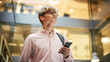 © Gorodenkoff - Portrait of Young Caucasian Man Feeling Confident as He Walks Smiling and Checking His Phone, in a Busy Office Hallway. He is Browsing the Internet on His Smartphone. Low Angle