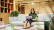 © Gorodenkoff - Portrait of Caucasian Creative Young Woman Working on a Laptop in Bright Casual Office. Female Client Coordinator Smiling When Receiving Positive Feedback From Clients. Employees Walking in Background