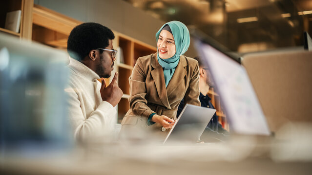 Fototapete - Two Coworkers Collaborating and Celebrating Their Accomplishment in a Modern Office. Muslim Female Trainer Using Laptop to Onboard a Black Male Customer Support Agent and Answering His Question.