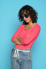 Wall Mural - Young exotic brunette woman in pink blouse and rope belt jeans posing with a sunglasses on a blue studio background