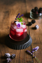 Bramble Cocktail With Violet Flower, Mint And Lavender On A Wood Backdrop