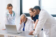 © dikushin - Serious medical team of multiethnic doctors talking together concentrated looking to laptop screen standing on background of window. Group of diverse practitioner doing online consultation via webcam