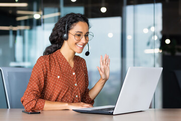 Happy hispanic woman working inside office, businesswoman with video call headset talking and advising customers remotely, tech support online store customer service, greeting with hand.