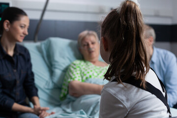 Wall Mural - Unrecognizable little girl chatting with lady bedridden suffering degenerative disease. Toddler accompanying grandmother before medical intervention by geriatrician clinic.