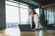 © DSMT - Smiling young woman in casual clothes and glasses, wearing a white jacket and laughing while sitting at a table with a laptop while working. Work in the office