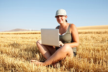 A Young Girl In A Blue Hat Smiles Toward Camera While Working On A Laptop Computer In The Middle Of A Fresh Cut Golden Wheat Field.