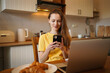 © Daryna  - Young attractive woman in yellow t-shirt typing on phone and working on laptop at home in modern kitchen. The concept of remote work and communication in social networks