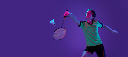 Wall Mural - Portrait of teen boy in uniform playing badminton, serving shuttlecock with racket isolated over blue purple background in neon ligth