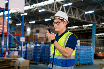 Asian male workers wearing reflective jackets and safety helmets use walkie-talkiescommunicating with colleagues inside warehouse distribution center to deliver supplies.Factory and logistics concept.