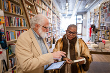 Bookshop Owner With Digital Tablet Helping Shopper With Book