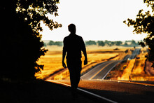 A Silhouetted Man Walking Along Side Of A Country Road.