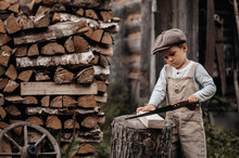 A Little Boy In A Cap Next To A Woodpile Saws Wood With A Saw In A Natural Landscape. Close-up. Gives The Picture An Authentic Mood. Vintage. Rustic.