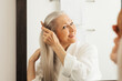 © Artem Varnitsin - Cheerful senior woman combing her hair looking at a mirror in bathroom