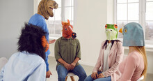 Group Of Funny People In Animal Disguise Having A Meeting In The Office. Team Of Young Men And Women Wearing Monkey, Horse, Frog And Pigeon Masks Sitting In A Circle And Listening To A Dinosaur Guy