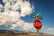 PANAMINT VALLEY, CA, USA. A Stop Sign And Two Street Signs In The Bottom Corner Of Image, With Desert Mountains On Horizon And A Big, Blue Sky With Puffy Clouds.