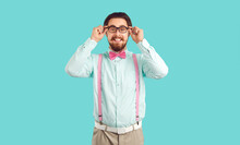 Young Caucasian Man Millennial With Funny Beard And Mustache Smiling Broadly Adjusts Glasses And Looks At Camera Dressed In White Shirt And Pink Bow Tie, Stands On Studio Background