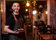 © alvaro - Waiter in uniform smiling at camera while holding a plate with food in a restaurant.