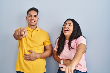 Poster - Young couple standing over isolated background laughing at you, pointing finger to the camera with hand over body, shame expression