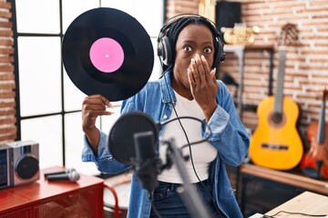 Wall Mural - Beautiful black woman holding vinyl record at music studio covering mouth with hand, shocked and afraid for mistake. surprised expression